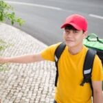 A cheerful delivery man in a red cap rings a doorbell in Portugal, wearing a yellow shirt and carrying an insulated bag.
