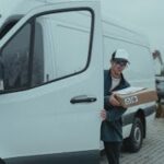 Delivery driver stepping out of van carrying a package on a cloudy day.