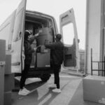 Two couriers loading cardboard boxes into a delivery van outdoors.